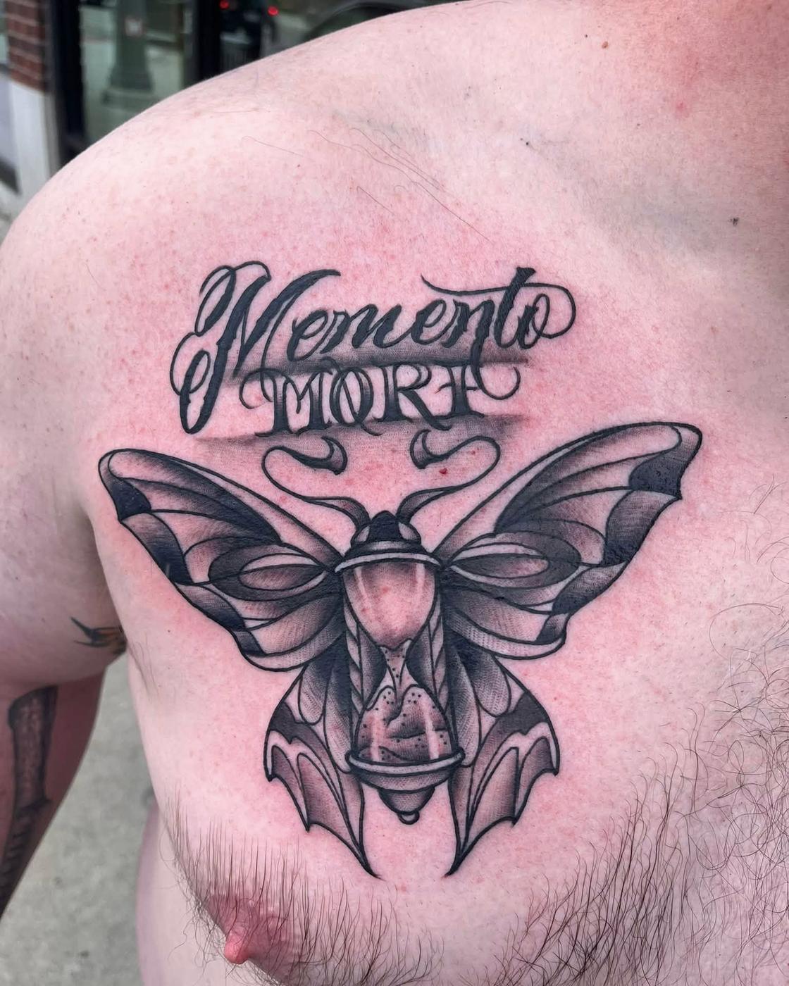 A black and gray moth beneath Memento Mori in script lettering, across a chest.