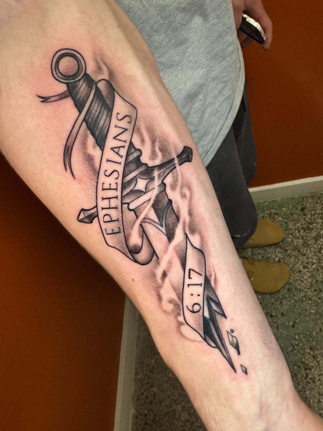 Black and grey sword with a lettered banner on a forearm.